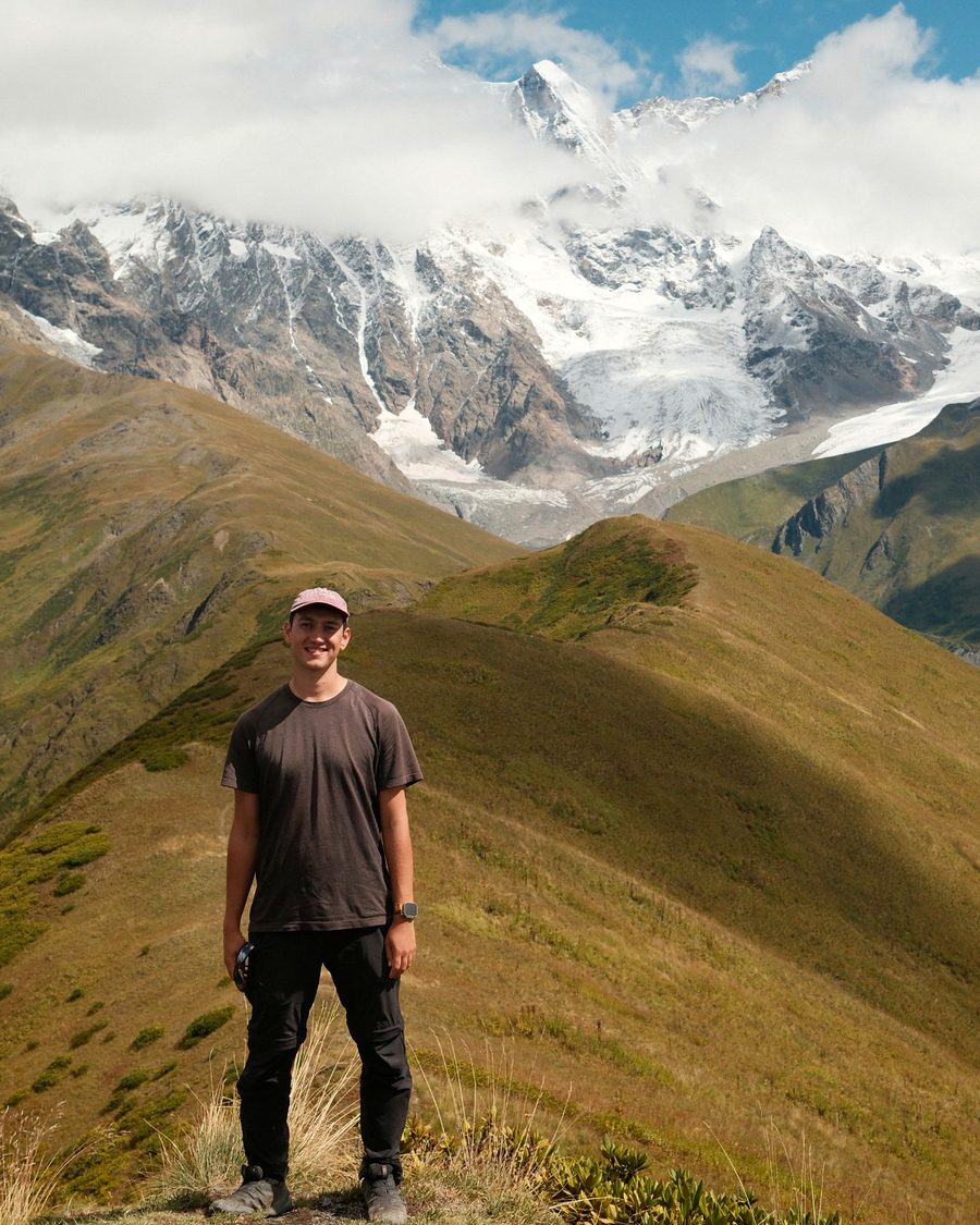Shastri, founder of Simple Web Design, standing on a grassy mountain ridge with snow-capped peaks behind him